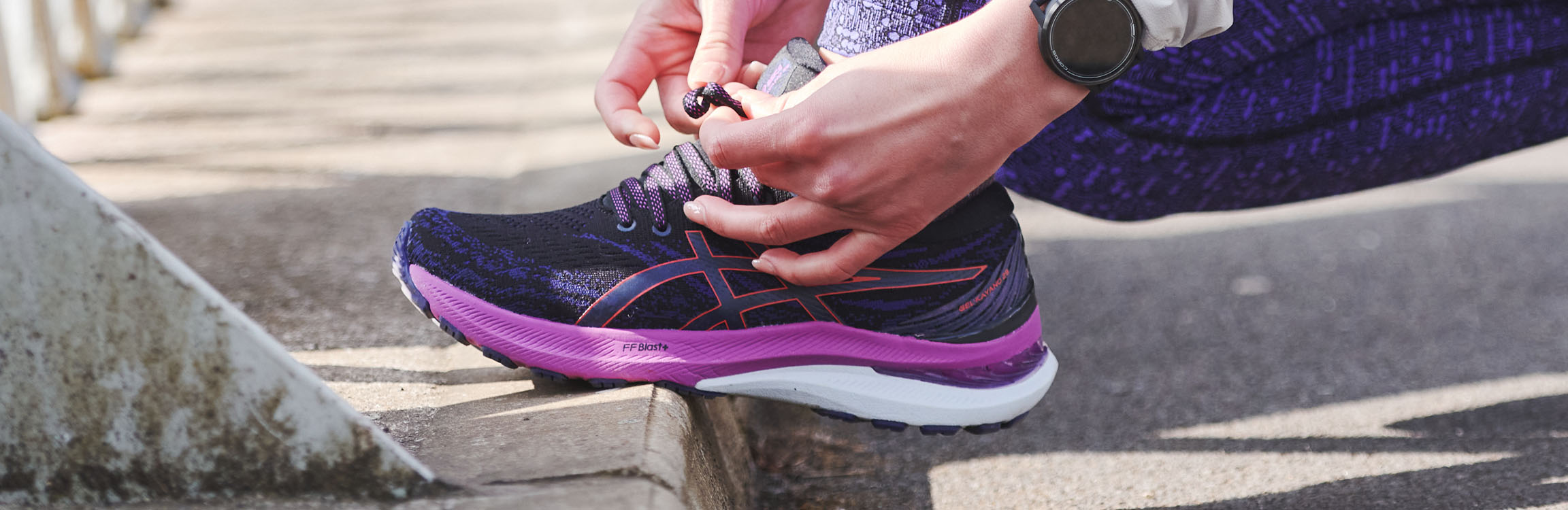 Person's hands are tieing shoe lases on Asics women's running shoe on a kerb.