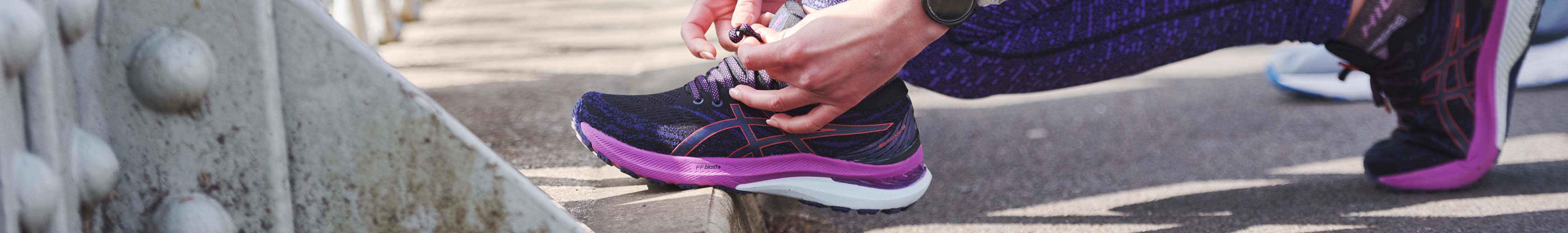 Person's hands are tieing shoe lases on Asics women's running shoe on a kerb.