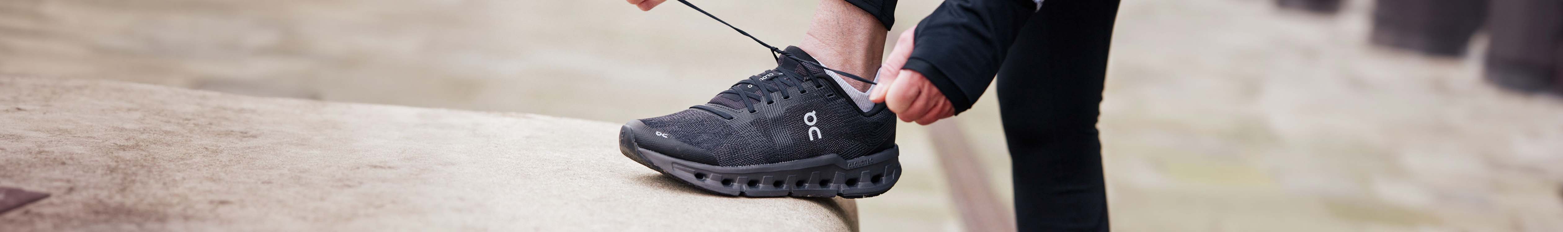 Shop men's road runnin shoes. A man tieing the lace on black ON shoe