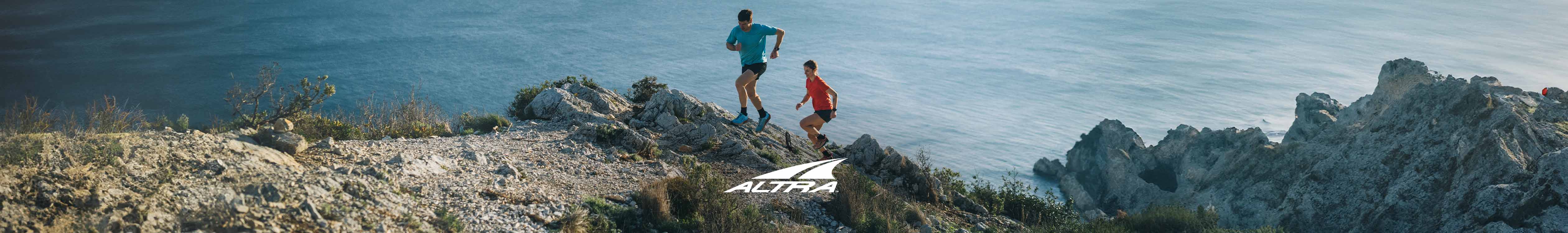 Shop Altra running shoes. Two people running on a rocky, mountainous trail with the sea in the background 