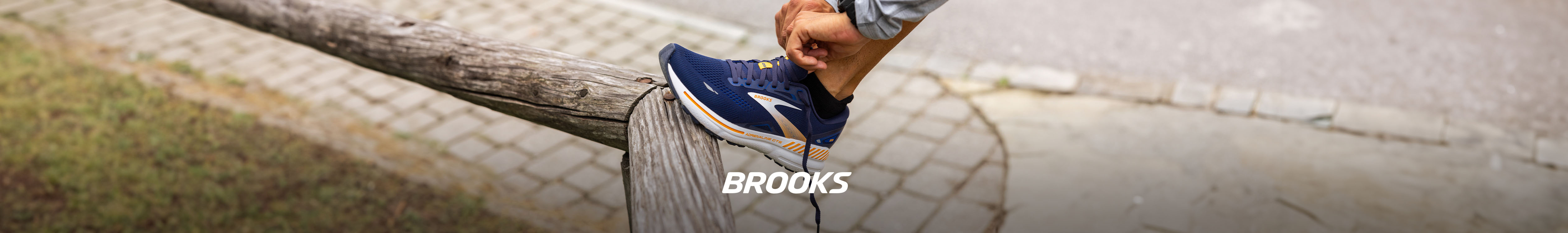 Shop Brooks. Person's foot in a blue Brooks shoe on a wooden railing. 