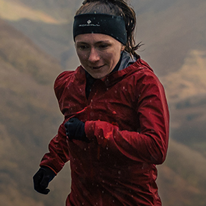 Shop hata & gloves new season. A woman in red jacket with black headband and gloves is running up the hill